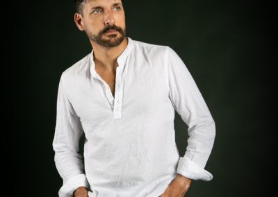 David Bello, en estudio, con fondo oscuro, camisa blanca y pantalón vaquero. Mirada lateral, plano americano.
