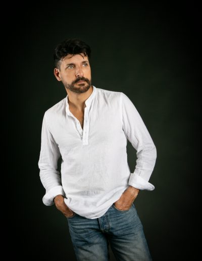 David Bello, en estudio, con fondo oscuro, camisa blanca y pantalón vaquero. Mirada lateral, plano americano.
